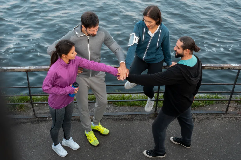 group of friends in workout clothes stacking hands by the water, showing that girl supportive relationships and healthy social energy