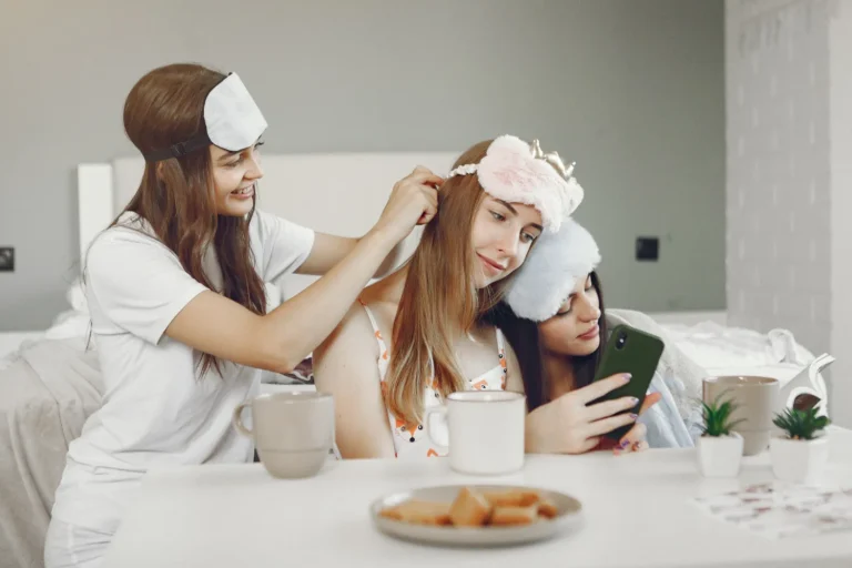 Three friends in sleepwear with fluffy sleep masks share a cozy pre-bed moment—braiding hair, scrolling a phone—mugs and snacks on the table.