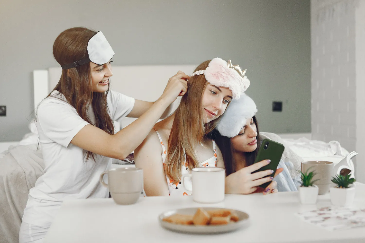 Three friends in sleepwear with fluffy sleep masks share a cozy pre-bed moment—braiding hair, scrolling a phone—mugs and snacks on the table.